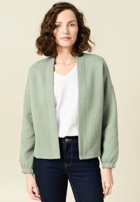 Cardigan trapuntato verde chiaro con una vestibilità ampia, scollatura a V e maniche arricciate, abbinato a una maglietta bianca e jeans scuri.