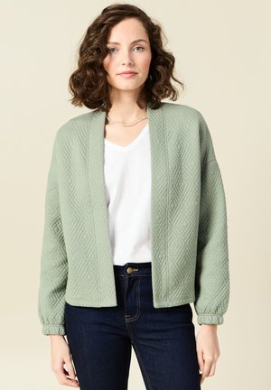 Hellgrüne, gesteppte Strickjacke mit lässiger Passform, V-Ausschnitt und gerafften Ärmeln, kombiniert mit einem weißen Oberteil und dunklen Jeans.
