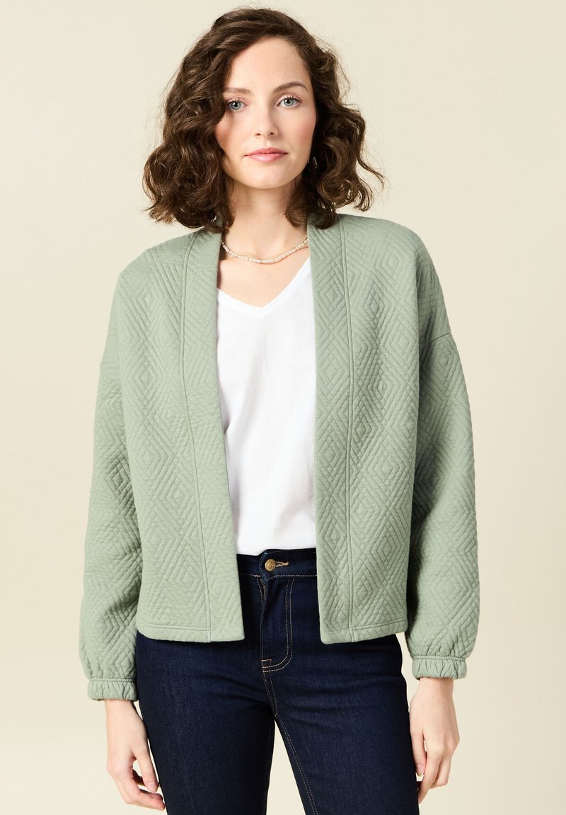 Cardigan trapuntato verde chiaro con una vestibilità ampia, scollatura a V e maniche arricciate, abbinato a una maglietta bianca e jeans scuri.