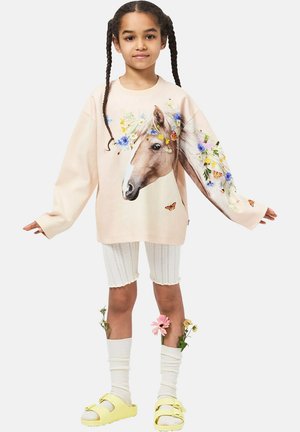 Vaaleanpunainen collegepaita, jossa on hevosen kuvio ja kukkaisia yksityiskohtia. Päällä valkoiset ribbineulosshortsit ja keltaiset sandaalit, joissa rusetit.