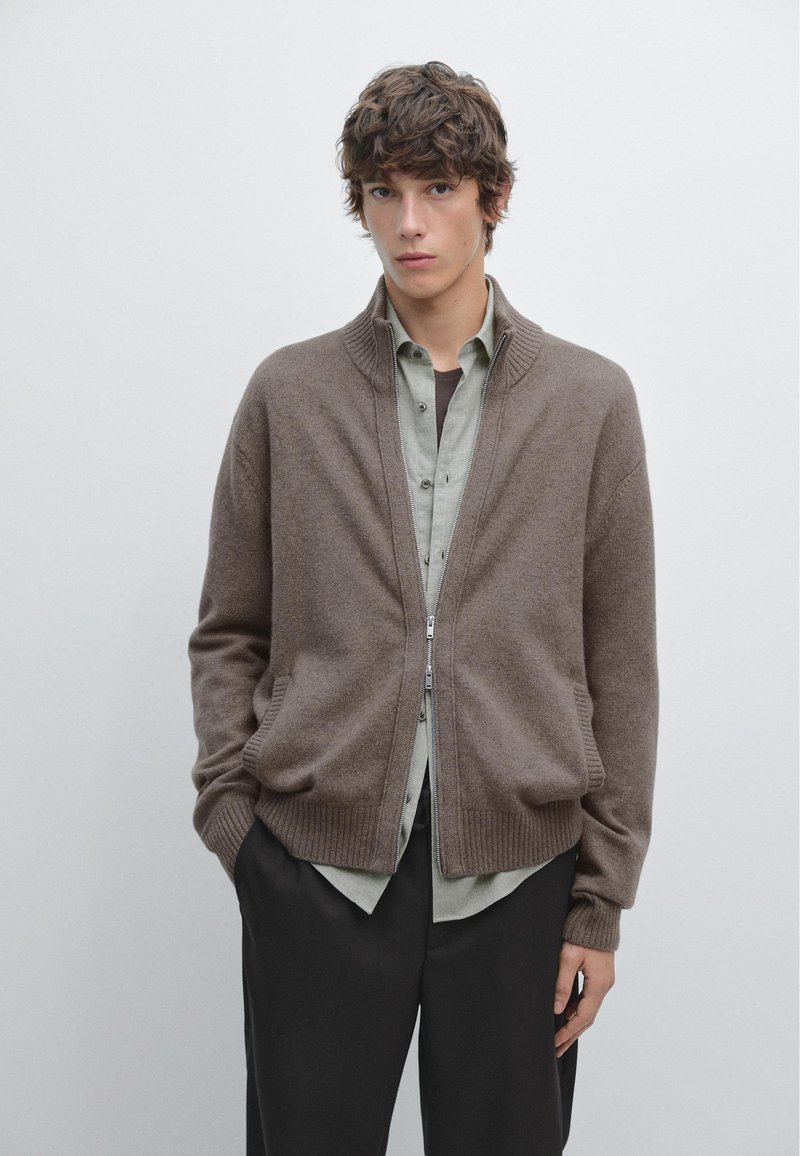 Brauner Reißverschluss-Cardigan mit geripptem Saum und Bündchen, aus weichem Material, getragen über einem hellgrünen Hemd mit Knöpfen und dunkler Hose.