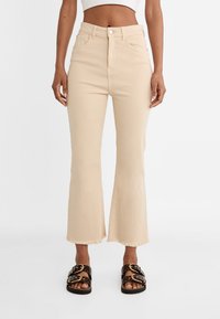 Mujer vestida con pantalones de campana cortos de talle alto en color beige con dobladillos deshilachados y sandalias negras con hebillas grandes.