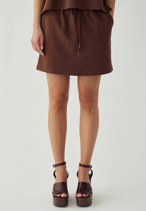 Žena oblečená v hnedej mini sukni a tmavohnedých platformových sandáloch na vysokom opätku s remienkami okolo členkov, stojaca pred jednoduchým pozadím.