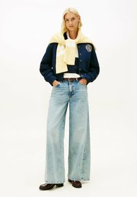 Mujer rubia con chaqueta azul marino abotonada, suéter amarillo sobre los hombros, vaqueros de pierna ancha de color azul claro y zapatos marrones, de pie con las manos en los bolsillos.