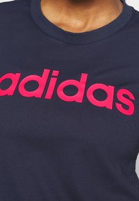 Marininblå bomullströja med rund hals och en framträdande rosa "adidas"-logotyp tryckt över bröstet. Slät textur, avslappnad design.