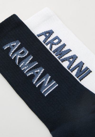Dos pares de calcetines: uno blanco con texto "ARMANI" en azul y negro, y otro azul oscuro con texto similar en un tono más claro. Tejido texturizado.