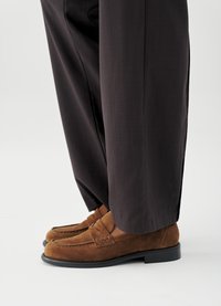 Καφέ σουέντ μοκασίνια με λείο δερμάτινο λουράκι, σε συνδυασμό με φαρδιά, σκούρα παντελόνια. Η σόλα είναι μαύρη και ελαφρώς ανυψωμένη.