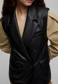 Gilet en similicuir noir à double boutonnage, avec des revers crantés et deux poches avant. Porté sur un haut beige clair à manches bouffantes.