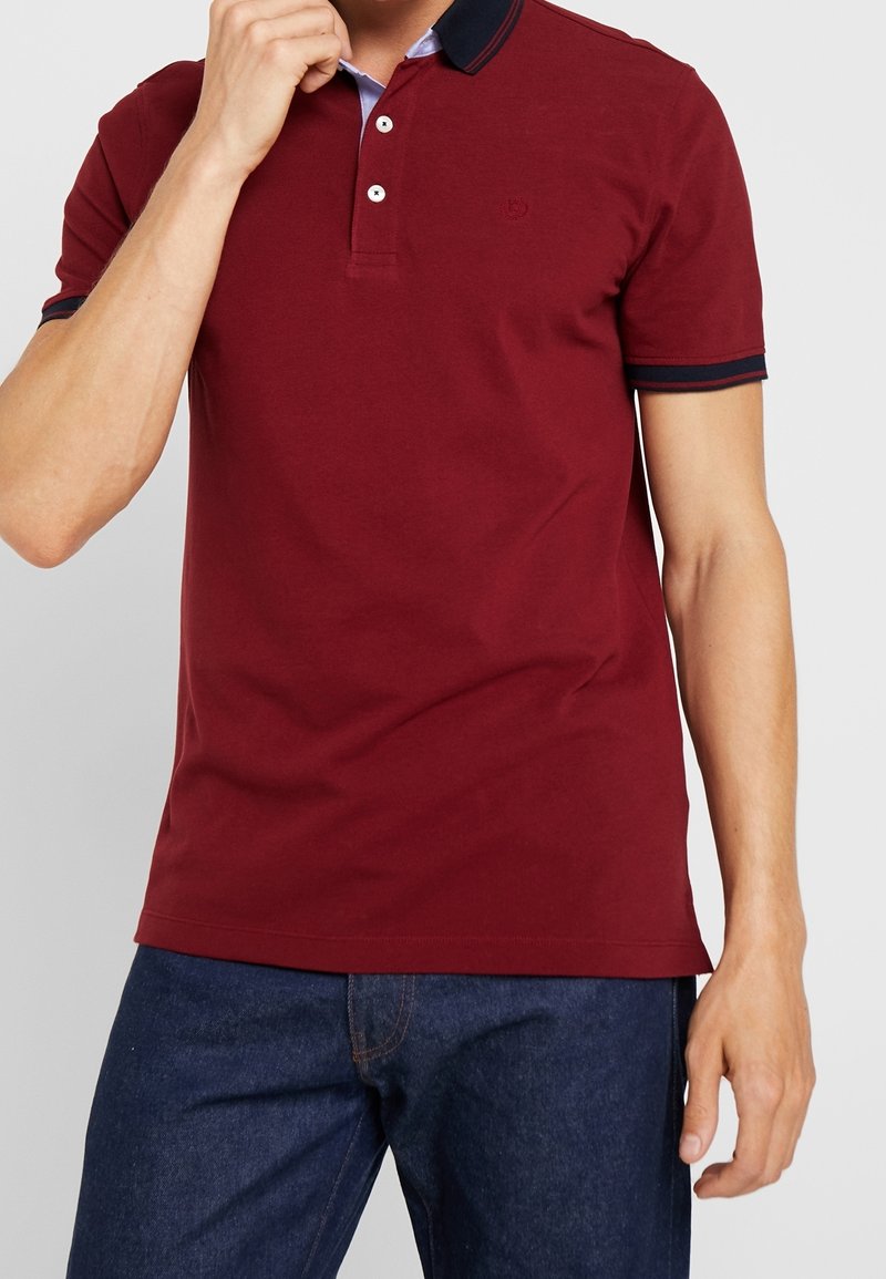 Homme portant un polo bordeaux avec un col et des bords de manches foncés, associé à un jean bleu foncé, main près du visage.