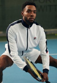 Weißer Reißverschlussjacke mit navy Ab accents, Fila-Logo auf der Brust, kombiniert mit weißen Shorts. Hält einen Tennisschläger, Hintergrund eines Hallenplatzes.