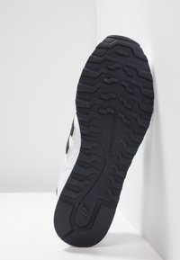 Suola in gomma nera di una sneaker bianca New Balance, con motivo del battistrada e logo del marchio su uno sfondo angolato bianco.