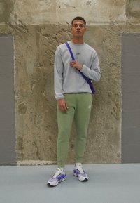 Grå hettegenser med en liten logo, grønne slim-fit sweatpants og flerfargede joggesko. Bakgrunnen har en teksturert betongvegg.