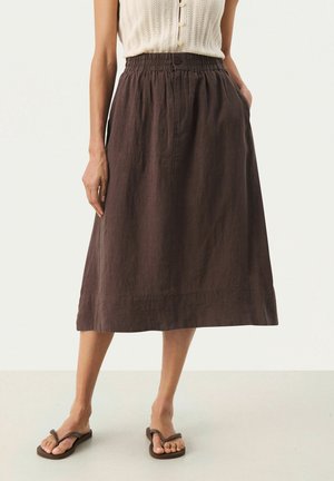 Femme portant une jupe midi marron avec une ceinture élastique, des poches latérales, un top en maille sans manches et des sandales marron de type tongs.