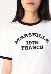 Hvit T-skjorte med svart ringhals som har teksten "MARSEILLE 1975 FRANKRIKE" i fet svart skrift på brystet.