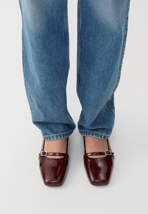 Voeten met bordeauxrode Mary Jane-schoenen met vierkante neus en blauwe spijkerbroek tegen een witte achtergrond.