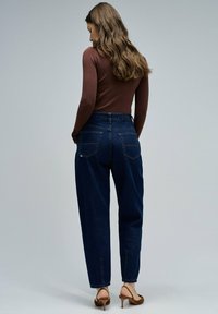 Vaqueros de tiro alto de color azul oscuro con un ajuste relajado, que cuentan con dos bolsillos traseros y dobladillo vuelto, combinados con una blusa marrón de manga larga.