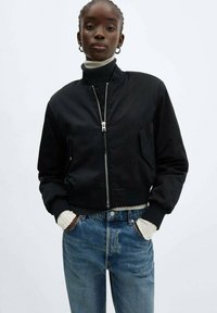 Schwarze, verkürzte Bomberjacke mit silberner Reißverschlussöffnung, seitlichen Taschen und gerippten Bündchen, kombiniert mit blauen Jeans.