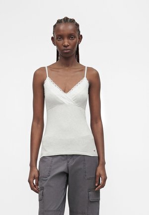 Vrouw met gevlochten haar die een wit kanten topje en grijze cargobroek draagt tegen een effen achtergrond.