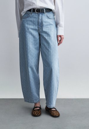 Világoskék, széles szárú farmer finom mintával, fekete öv, fehér hosszú ujjú ing és leopárdmintás Mary Jane cipők szürke padlón.