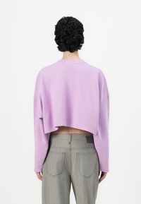 Lyhennetty, ylisuurta hupparia pastellivioletissa värissä, valmistettu pehmeästä kankaasta. Varustettu leveillä hihoilla ja pyöreällä kaula-aukolla, yhdistettyinä korkeavyötäröisiin harmaisiin housuihin.