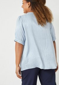 Hellbluse mit strukturiertem Muster, kurzen Ärmeln mit gerafften Bündchen, fließender Passform und abgerundetem Saum, getragen mit dunklen Hosen.