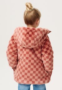 Geruite roze en crèmekleurige fleecejas met een capuchon, met een zachte textuur en geribde manchetten, gedragen over blauwe jeans.