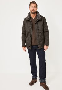 Dunkelgrüne Jacke mit einem Kunstpelzkragen, Reißverschluss und Knopfverschluss, Brusttaschen, kombiniert mit einem braunen Pullover und dunklen Jeans. Braune Schuhe runden den Look ab.