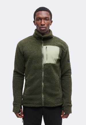 Homme portant une veste polaire verte zippée avec une poche poitrine zippée vert clair, debout devant un fond clair uni.