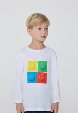 Blondýnský chlapec v bílém tričku s dlouhým rukávem, na kterém jsou čtyři barevné potisky LEGO kostek v barvách žlutá, zelená, modrá a červená, dívající se stranou.