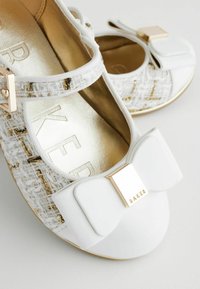 Weiße Ballettschuhe aus strukturiertem Stoff mit goldenen Akzenten, ausgestattet mit einer großen weißen Schleife und einem goldenen Logodetail auf der Vorderseite.