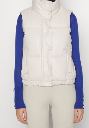 Chaleco beige acolchado con cuello alto, cierre frontal abotonado y bolsillos laterales. Lleva encima de una camisa de manga larga azul con puños adornados.