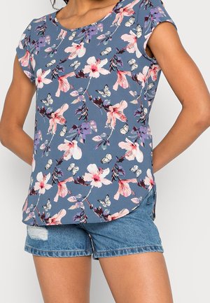 Blomstermønster top i lyseblå med pink og lilla blomster samt hvide sommerfugle; korte ærmer og rund kant. Sat sammen med denimshorts.