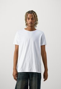 Camiseta branca de manga curta com decote redondo e ajuste descontraído. O tecido parece suave com uma textura leve. Utilizador vestido com jeans escuros.