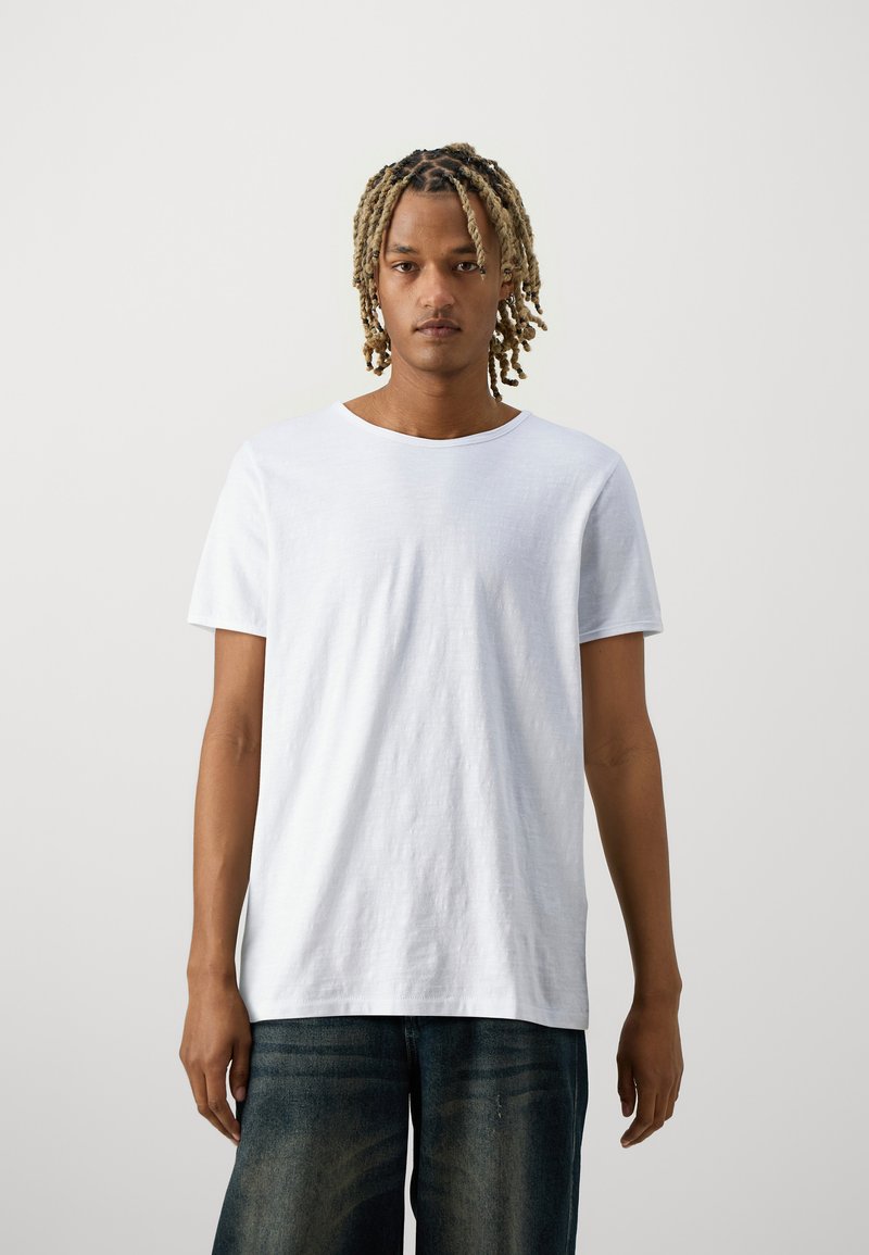 Camiseta branca de manga curta com decote redondo e ajuste descontraído. O tecido parece suave com uma textura leve. Utilizador vestido com jeans escuros.