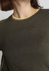 Olijfgroene T-shirt met een ronde halslijn, contrasterende gele afwerking en een gestructureerde stof. Korte mouwen met subtiele stikdetails.