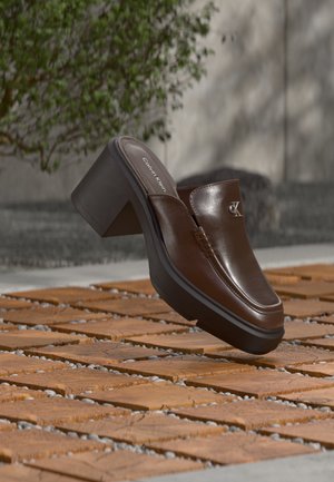 Braune Leder-Calvin Klein Absatzpantolette, die über einem Holzboden mit verschwommenem grünem Pflanzen- und Wandhintergrund schwebt.