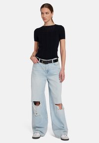 Top nero a costine con maniche corte, abbinato a jeans larghi chiari strappati e cintura nera; modello che indossa sneaker bianche e grigie.