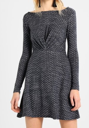 Mujer con un vestido de manga larga gris oscuro con pequeños lunares blancos y un detalle fruncido en la cintura.