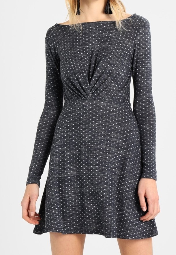 Femme portant une robe à manches longues gris foncé avec de petits pois blancs et un détail froncé à la taille.