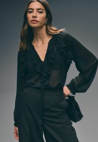 Sort gennemsigtig bluse med flæse detaljer omkring halsudskæringen og manchetterne, parret med højtaljede sorte bukser. Modellen holder en lille sort taske.