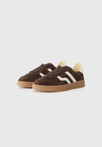 Sneaker in suede marrone con strisce bianche e dettagli beige. Suola in gomma, design con lacci e punta arrotondata. Estetica minimalista.