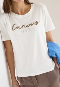 Femme portant un t-shirt blanc avec le mot "Curious" en sequins et un petit texte, tenant un pull bleu, debout contre un mur clair.