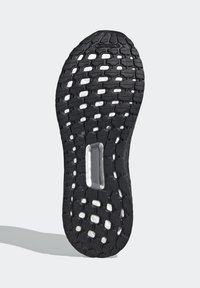 Semelle de chaussure en caoutchouc noir avec mousse blanche visible à travers des motifs découpés et un coussinet rectangulaire central, sur fond gris clair.
