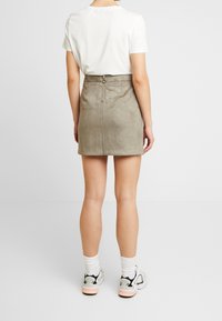 Suede mini sukně světle olivové barvy, s krytým zipem na zadní straně, přiléhavým střihem a hladkým povrchem, spárovaná s bílými teniskami.