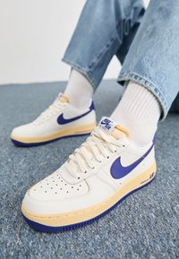 Personne portant des baskets Nike Air Force 1 blanches avec des swooshes bleus, des semelles intermédiaires crème, des chaussettes blanches et un jean bleu clair sur un tapis bleu.