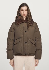 Giovane donna che indossa un giubbotto imbottito marrone con bottoni grandi, tasche a soffietto e un colletto in pelliccia scura, in piedi contro uno sfondo semplice.