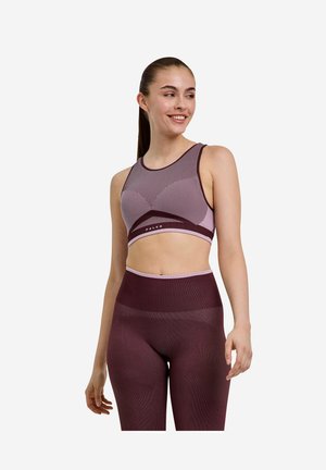 Junge Frau trägt passende lila hoch taillierte Leggings und gemustertes Crop-Top, lächelt und schaut zur Seite vor weißem Hintergrund.