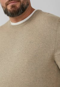 Beige gestrickter Pullover mit Rundhalsausschnitt, der eine subtile Textur und ein geprägtes Logo auf der Brust hat. Der innere Kragen ist weiß.