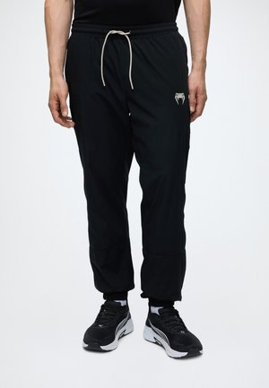 Hombre con pantalones jogger negros con cordón blanco, zapatos deportivos negros y calcetines blancos sobre un fondo blanco liso.
