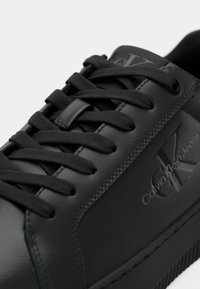 Zapatillas negras de cuero con cordones planos negros, con una superficie texturizada y logo "CK" en relieve en el lateral. Suela de goma.